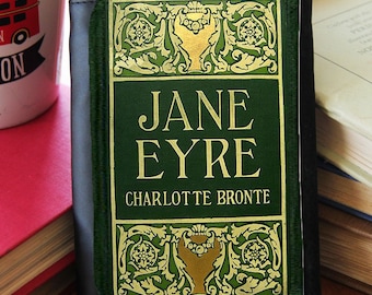 Könnte beinhalten: Eine schwarze Lederbörse mit grünem und goldenem Design. Die Vorderseite der Börse zeigt den Titel "Jane Eyre" von Charlotte Bronte.