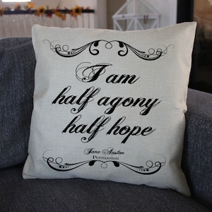 May include: A square beige throw pillow featuring black text that reads, I am half agony half hope, followed by Jane Austen and Persuasion. The design includes decorative black scrollwork borders at the top and bottom of the quote.