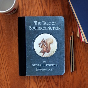 Op de afbeelding: Een blauw notitieboek met een zwarte rug. De omslag heeft een vintage illustratie van een eekhoorn die een noot vasthoudt, met de tekst "The Tale of Squirrel Nutkin" en "By Beatrix Potter, F. Warne & Co."