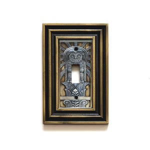 May include: A rectangular light switch plate featuring a metallic silver owl perched atop a decorative column, set against a sunburst background. The plate is framed by a dark bronze-colored border with stepped, tiered edges and two screw holes.