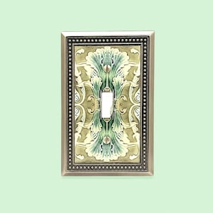 May include: A rectangular light switch plate featuring a metallic silver border with a beaded edge. The center panel displays a symmetrical, embossed pattern in shades of sage green, cream, and white, with a central vertical toggle opening.
