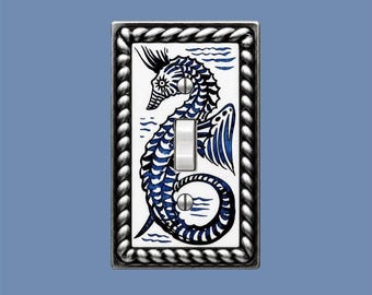 May include: A rectangular light switch plate featuring a white ceramic center with a blue seahorse illustration and wavy water lines. The plate is framed by a metallic, silver-toned rope-style border and includes two screws and a central toggle.