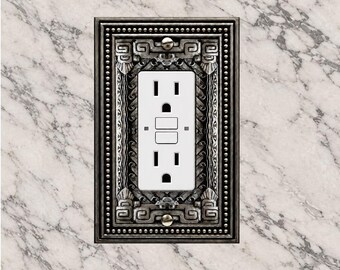 May include: A rectangular wall plate with a dark metallic finish and intricate embossed geometric patterns surrounding a white GFCI electrical outlet. The plate features a beaded border and is secured with two visible metal screws.