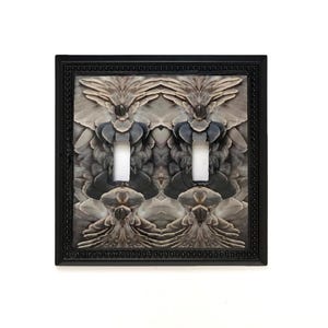 May include: A double toggle light switch plate featuring a black frame with a beaded edge. The center panel displays a symmetrical, mirrored pattern of grey, brown, and black feathers, with two rectangular cutouts for switches and two black screws.