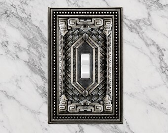 May include: A rectangular light switch wall plate featuring an intricate, dark metallic finish with a raised, geometric, and repeating pattern. The plate has a beaded border and a central white toggle switch secured by two visible metal screws.