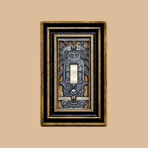 May include: A rectangular light switch plate featuring a central silver-toned owl relief design with a sunburst pattern. The plate has a dark, distressed gold and black border with two visible screws securing the switch mechanism in the center.
