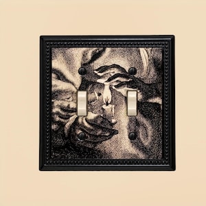 May include: A double light switch cover plate featuring a black and white etched illustration of hands lighting a candle. The square plate has a decorative beaded border and two vertical toggle switches in the center, secured by four black screws.