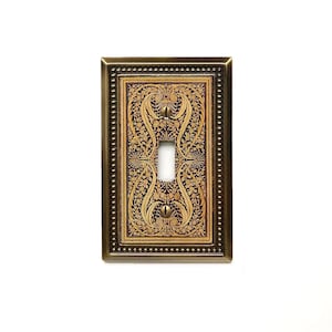 May include: A rectangular light switch wall plate with a metallic bronze finish and a beaded border. The center features an intricate, dark brown and gold floral scrollwork pattern with a vertical toggle switch opening and two matching screws.
