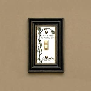 May include: A rectangular light switch plate with a dark bronze-toned frame and a white center panel. The panel features green botanical illustrations and the black text IN THE GARDEN and DREAMS, with a central toggle switch and two screws.