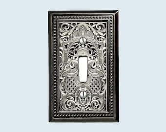 May include: A rectangular light switch plate featuring a dark metallic finish with intricate silver-toned scrollwork and lattice patterns. The plate has a beaded border and two visible screws securing it to the wall, with a central toggle opening.