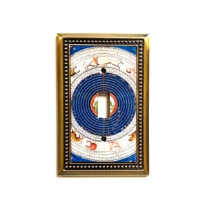 May include: A decorative light switch cover featuring a celestial design. The cover has a brass-colored frame with a detailed border. The center design includes a blue circle with concentric rings and zodiac symbols. The switch opening is centered.
