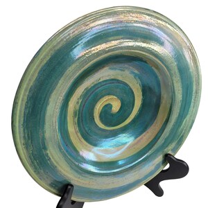 Puede incluir: Un plato grande y redondo de cerámica con un esmalte verde y dorado en espiral. El plato está sobre un soporte negro.