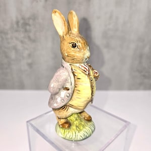 May include: A ceramic figurine of a rabbit, wearing a light yellow vest, a pink jacket, and a red and white striped tie. The rabbit stands on a patch of green grass. The figurine has detailed facial features and is set on a clear display stand.