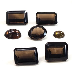 May include: A collection of six loose smoky quartz gemstones in various shapes and cuts. The set includes four rectangular faceted stones, one oval faceted stone, and one small smooth cabochon, all displaying a translucent dark brown color.