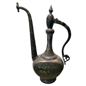 May include: Antique metal pitcher with a long, curved spout and a handle. The pitcher has a textured surface and a decorative finial on the lid. The overall color is a dark, aged metal, suggesting it is an old piece.