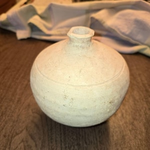 May include: A small, round, off-white ceramic vase with a textured, matte surface. The vessel features a narrow, circular neck opening and a subtle horizontal incised line encircling the upper portion of the body, showcasing a rustic, hand-formed appearance.