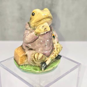 May include: A ceramic figurine of a frog in a seated position, wearing a pink coat. The frog has yellow skin, black eyes, and is holding a yellow object. It sits on a green base with a brown square. The figurine is a decorative item.