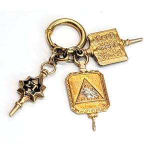 May include: A gold-toned keychain with three decorative charms. One charm features the Greek letters ΣΤ, another has a triangular emblem, and the third has text that reads "Who Studies in American Universities and Colleges."