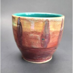 Puede incluir: Una taza de cerámica hecha a mano con forma redondeada. La taza presenta una vibrante paleta de colores, incluyendo tonos de rojo, naranja y amarillo, con vetas verticales de color púrpura oscuro. El borde es de color turquesa brillante. La taza es adecuada para bebidas.