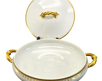 May include: White porcelain serving bowl with a lid, featuring gold trim and handles. The lid is off to the side, revealing the bowl's interior. The bowl has a decorative gold pattern around the base and on the lid's handle.