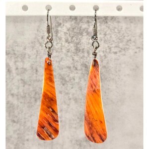May include: A pair of dangle earrings featuring elongated, teardrop-shaped pendants made of polished shell material. The pendants display a vibrant orange color with dark brown, streaky, natural patterns. Each earring hangs from a silver-toned metal hook.