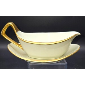 May include: Cream-colored gravy boat with a matching saucer. The boat has a gold-colored handle and trim. The gravy boat is sitting on a clear surface against a black background. The gravy boat is a classic piece of tableware.