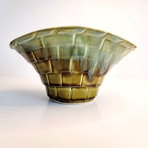 May include: A green ceramic planter with a woven pattern. The planter has a wide, flared opening and a narrow base.