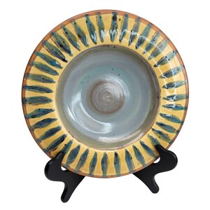 Puede incluir: Un plato de cerámica hecho a mano con un borde amarillo y patrones de hojas azules y amarillas. El centro del plato es de color azul verdoso claro. El plato se exhibe en un soporte negro.