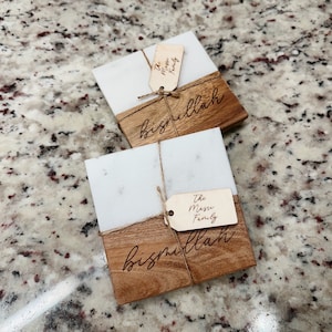 May include: Two white marble and wood coasters with the word "bismillah" engraved on the wood. Each coaster has a small wooden tag attached with the text "The Masse Family" on one and "The Mom Family" on the other.