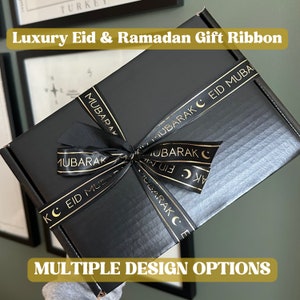 May include: Black gift box with a black ribbon tied in a bow. The ribbon has gold lettering that says "Eid Mubarak" and "Eid Mubarak C".