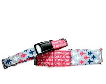 May include: A red and blue dog collar with a white background and a repeating pattern of fighter jets. The collar has the text "Before Flight Remove Before Flight" printed on it.