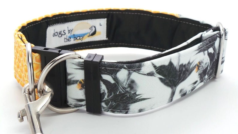 May include: A black and white dog collar with a yellow honeycomb pattern on one side and a black and white floral pattern on the other side. The collar has a black buckle and a silver D-ring. The collar is labeled "dogs by the bay".