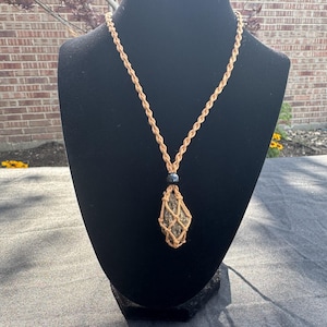 May include: A necklace featuring a rough, dark-colored stone pendant encased in a tan-colored macrame cord netting. The pendant is topped with a single, smooth, round black bead, and the necklace hangs from a matching twisted tan cord chain.