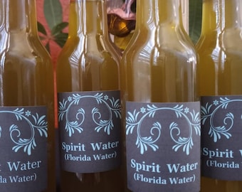 May include: Four glass bottles filled with a light brown liquid. Each bottle has a black label with white text and a blue floral design. The text on the labels reads "Spirit Water (Florida Water)".