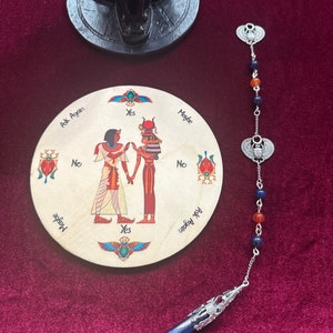 May include: A wooden planchette with Egyptian figures and text, alongside a pendulum with a blue gemstone and silver accents. The planchette has "Yes", "No", and "Maybe" options. A black cat statue is in the background.