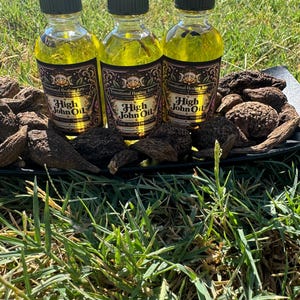 May include: Three clear glass bottles filled with yellow oil, each featuring a black cap and a label that reads Transitional Healing Arts High John Oil Power Success Protection, surrounded by several dark brown, textured, dried roots.