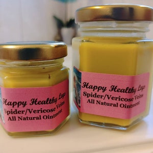 Puede incluir: Dos pequeños frascos de vidrio con tapas doradas llenos de un ungüento amarillo. Ambos frascos tienen etiquetas rosadas con texto negro que dice "Happy Healthy Legs Spider/Vericose Veins All Natural Ointment".