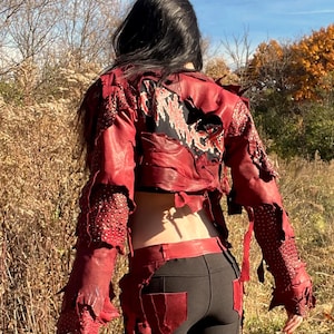 May include: Red leather jacket with distressed details, paired with black leggings and matching red boots. The jacket features a graphic design on the back. The outfit is suitable for cosplay or alternative fashion.