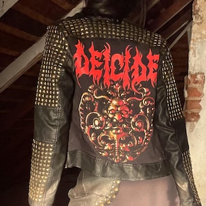 May include: Black leather jacket with gold studs on the shoulders and sleeves. The back of the jacket features a graphic with the word "DEICIDE" in red and a detailed illustration. The jacket is part of a gothic or heavy metal fashion style.