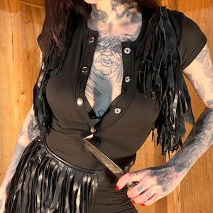 May include: Black outfit featuring a short-sleeved top with silver buttons and a fringed vest. A matching fringed belt and black leggings complete the ensemble. A knife is held at the waist. The outfit is a dark, edgy style.