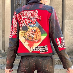 May include: A red and black leather jacket with the words "Cannibal Corpse" and "Hammer Smashed Face" printed on the back. The jacket has a graphic design on the back and sleeves, and is paired with matching pants.