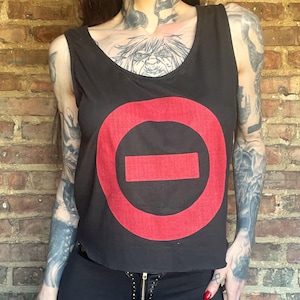 May include: Black tank top with a large red circle and horizontal rectangle graphic. The top is worn with black pants. The background is a brick wall.