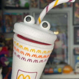 May include: A white plush cup with large googly eyes and a striped straw. The cup features the McDonald's logo and repeating golden arches. The rim is red, and the straw has yellow and purple stripes.