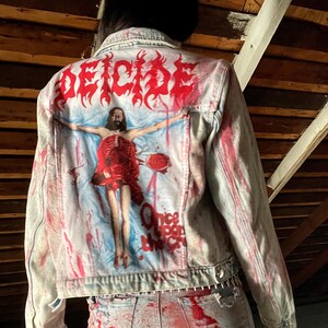 May include: Light-wash denim jacket and jeans set, splattered with red paint. The jacket features a graphic on the back with the word "DEICIDE" in red, and a religious-themed illustration. The jeans have distressed details.