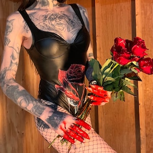May include: A black bodysuit with a sweetheart neckline and a rose graphic. The person is holding a bouquet of red roses. The hands are painted red, and the person is wearing fishnet stockings. The background is a wooden wall.
