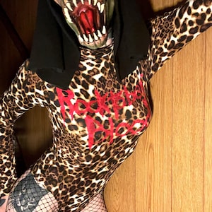 May include: A leopard print bodysuit with the red text "Rockstar" across the chest. The person is wearing a monster mask with a black hood and fishnet stockings. The background is a wooden wall.