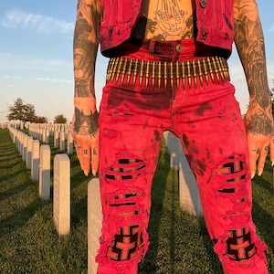 May include: A person wearing a red denim vest and distressed red jeans with black cross patches. The jeans have large rips and a belt made of bullet casings. The background shows a cemetery with white headstones.