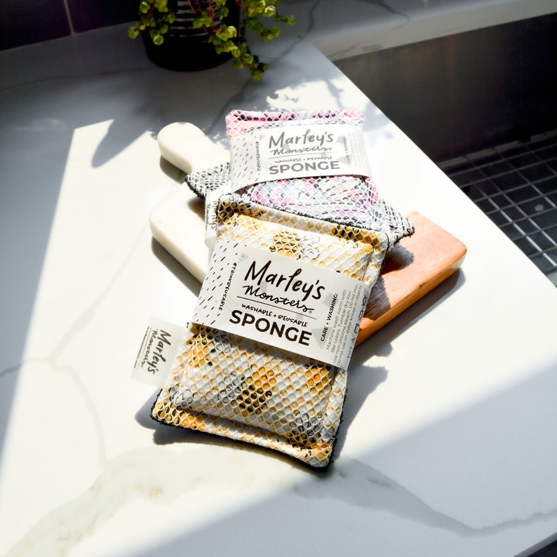 May include: Three yellow, pink, and gray reusable kitchen sponges with a honeycomb pattern. The sponges are labeled "Marley's Monsters" and say "Washable + Reusable Sponge".