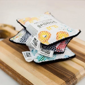 May include: Three colorful kitchen sponges stacked on a wooden cutting board. The top sponge is white with a colorful pattern and the text "Multi-Purpose Sponge" and "SPONGE". The bottom sponge is green with a blue and white pattern. The middle sponge is red with a black and white pattern.