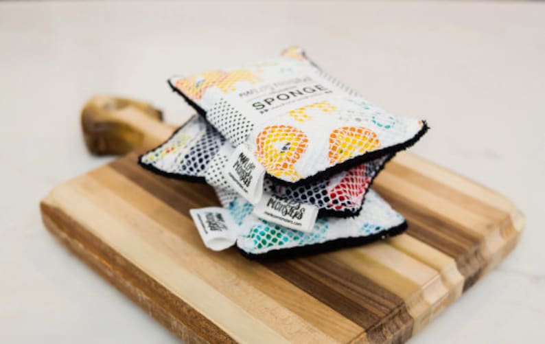 May include: Three colorful kitchen sponges stacked on a wooden cutting board. The top sponge is white with a colorful pattern and the text "Multi-Purpose Sponge" and "SPONGE". The bottom sponge is green with a blue and white pattern. The middle sponge is red with a black and white pattern.